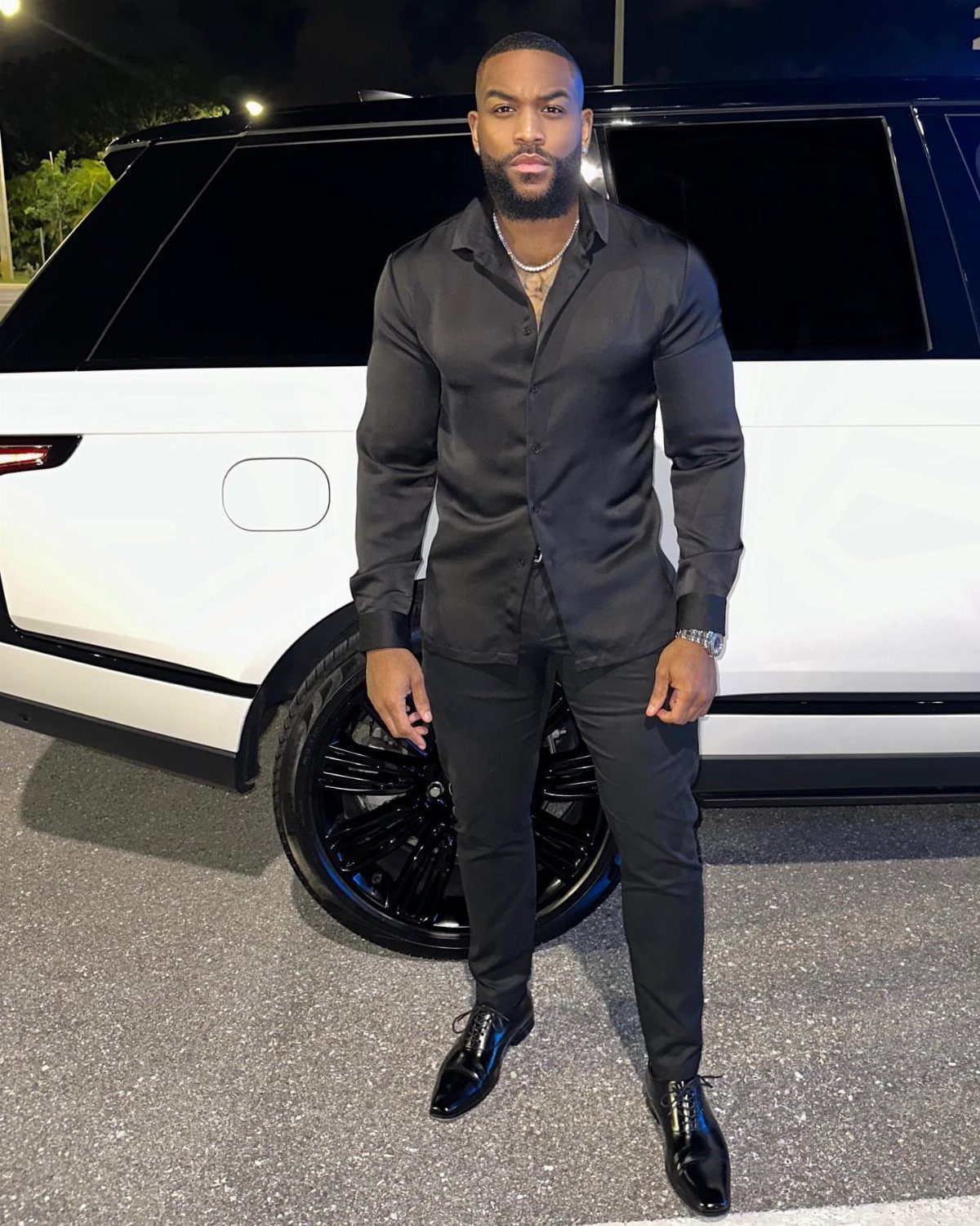 Man in all black cocktail attire standing in front of a white vehicle at night.