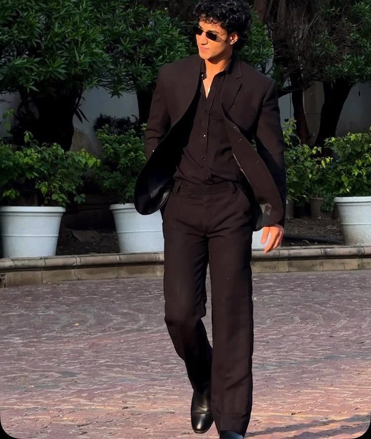 A man in an all-black suit and sunglasses walking through an outdoor courtyard.