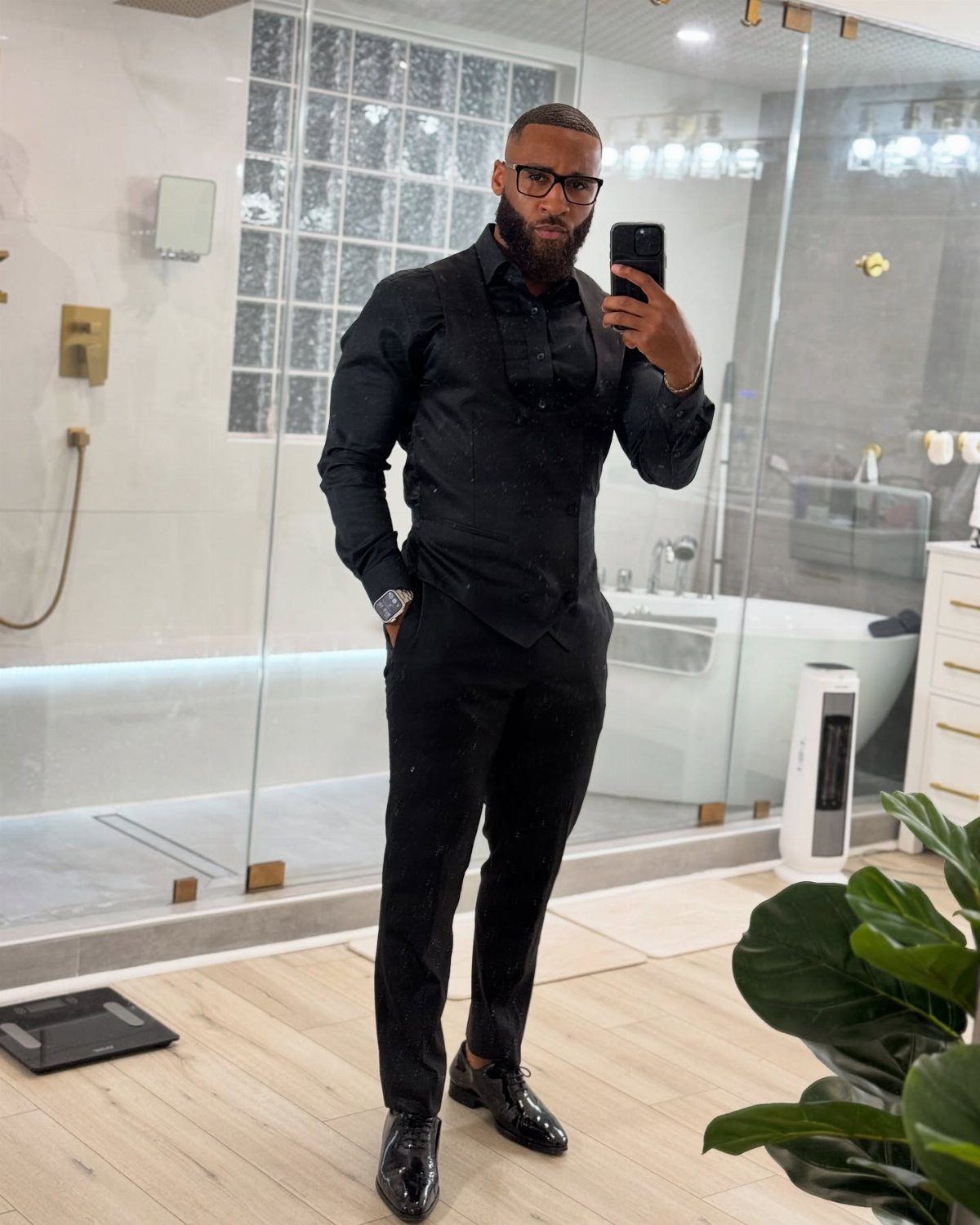 Man in all black cocktail attire posing for a mirror selfie in a modern bathroom.