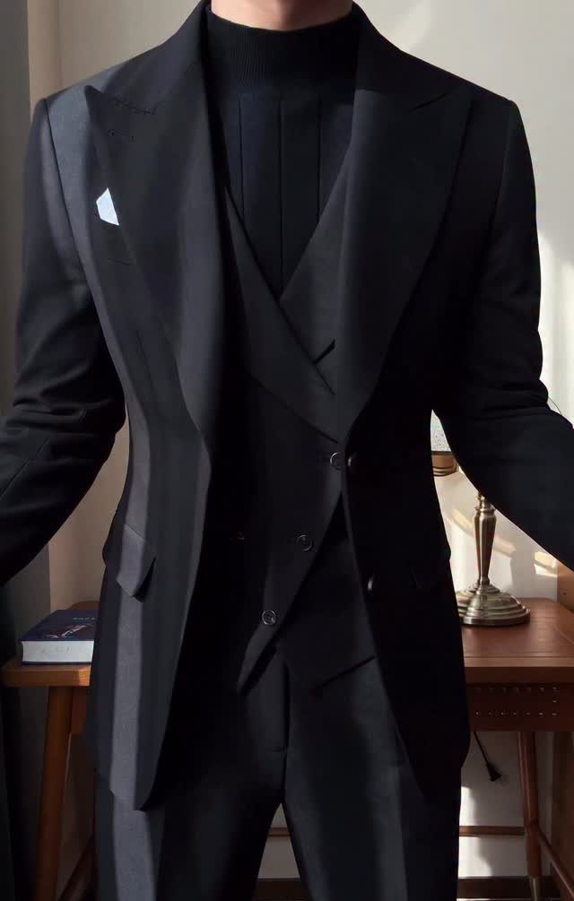 Man wearing a black blazer, waistcoat, and turtleneck sweater in a dimly lit room.