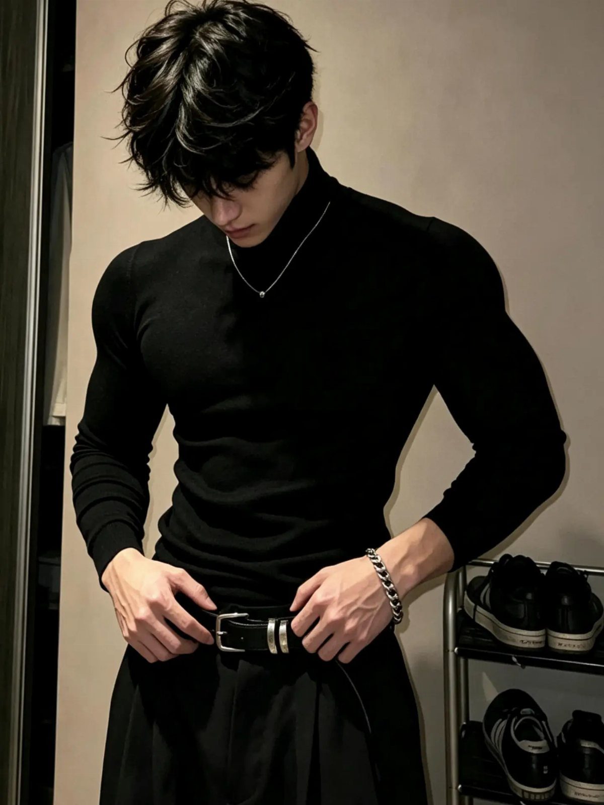 Man in a black turtleneck and trousers adjusting his belt while wearing a silver chain.