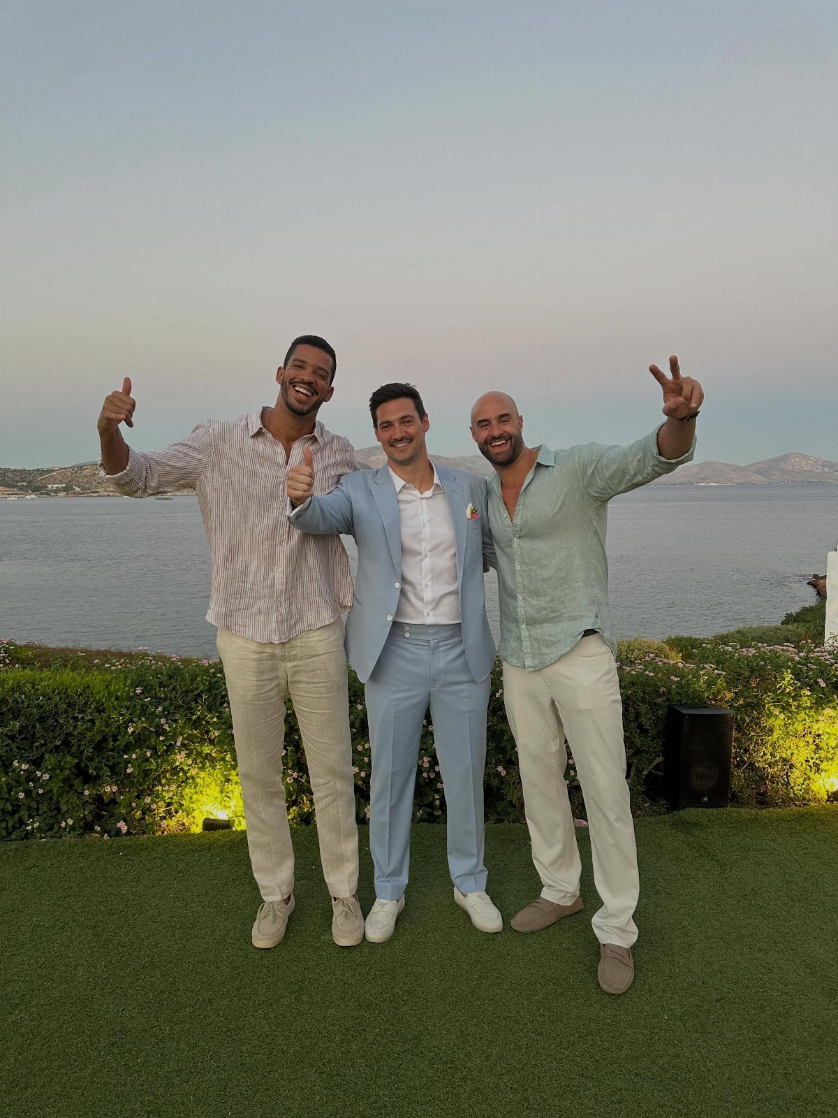 Three men smiling and posing in light-colored summer outfits by the sea at dusk.