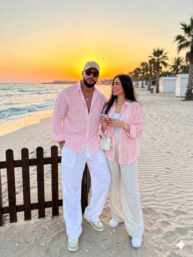 Man in pink linen shirt and white trousers posing with a woman on a beach at sunset.