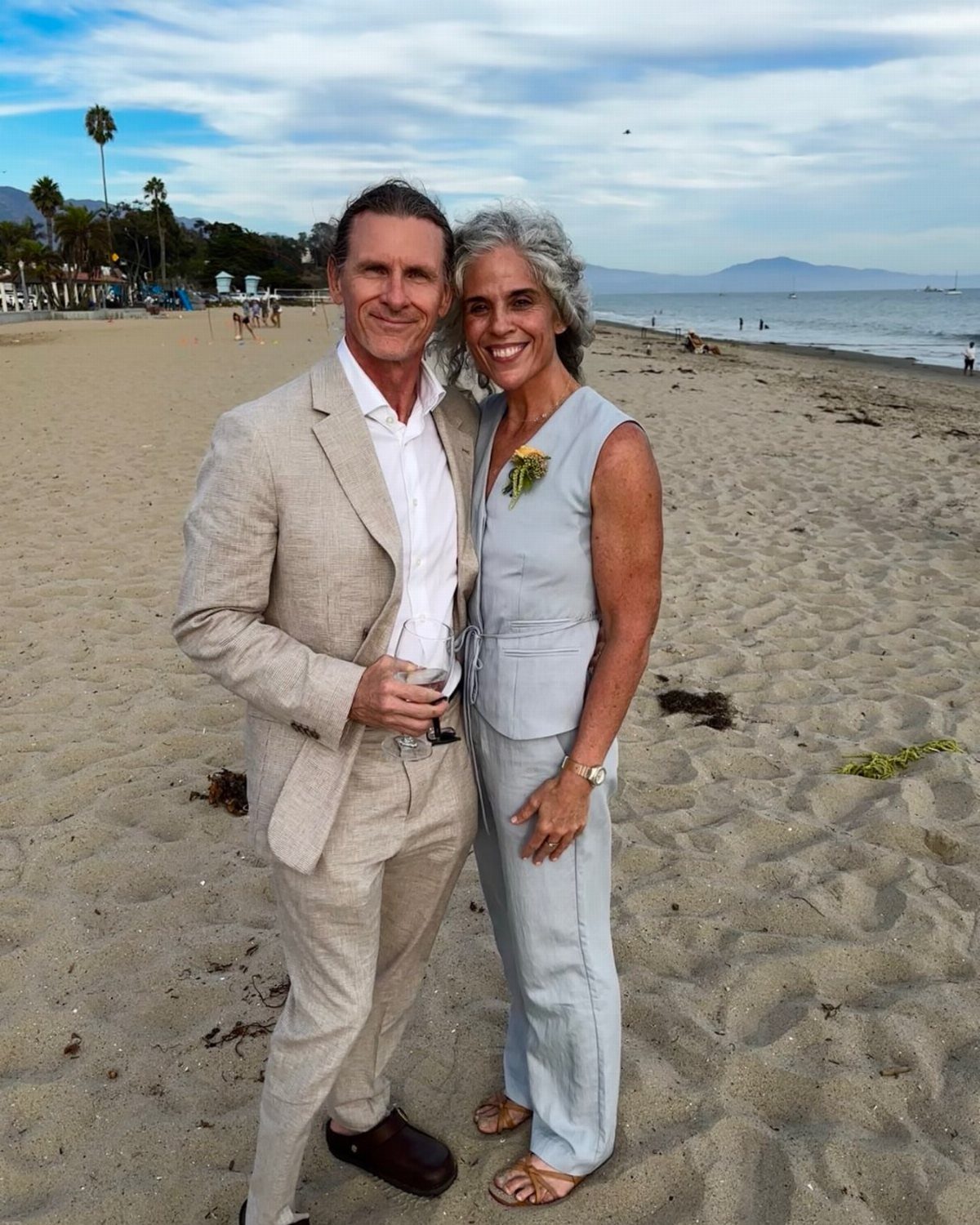 Man in a beige linen suit and woman in a blue jumpsuit posing on a beach.