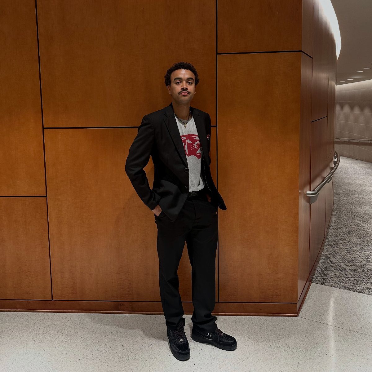 Man in a black blazer and white graphic tee posing against a wooden wall.