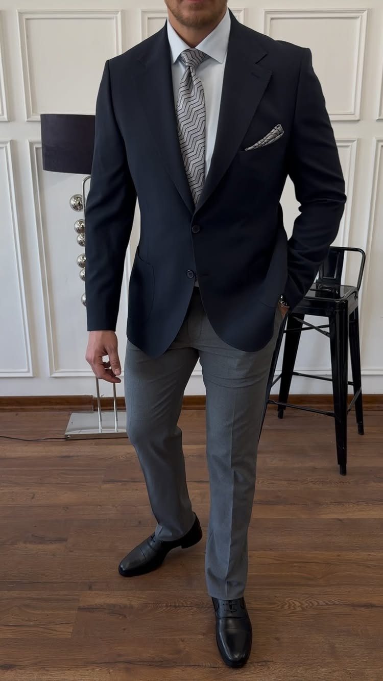 Man in a dark blazer, white shirt, patterned tie, and grey trousers standing indoors.