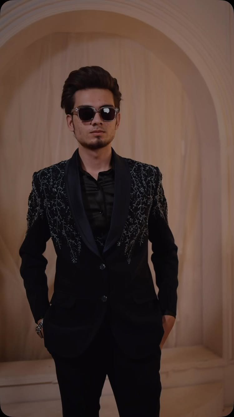Man in an embroidered black blazer and sunglasses standing against an arched wall.
