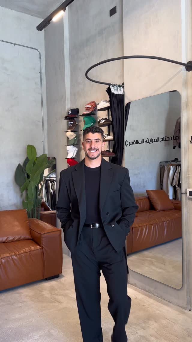 A smiling man wearing a black blazer, black sweater, and black trousers standing in a room.