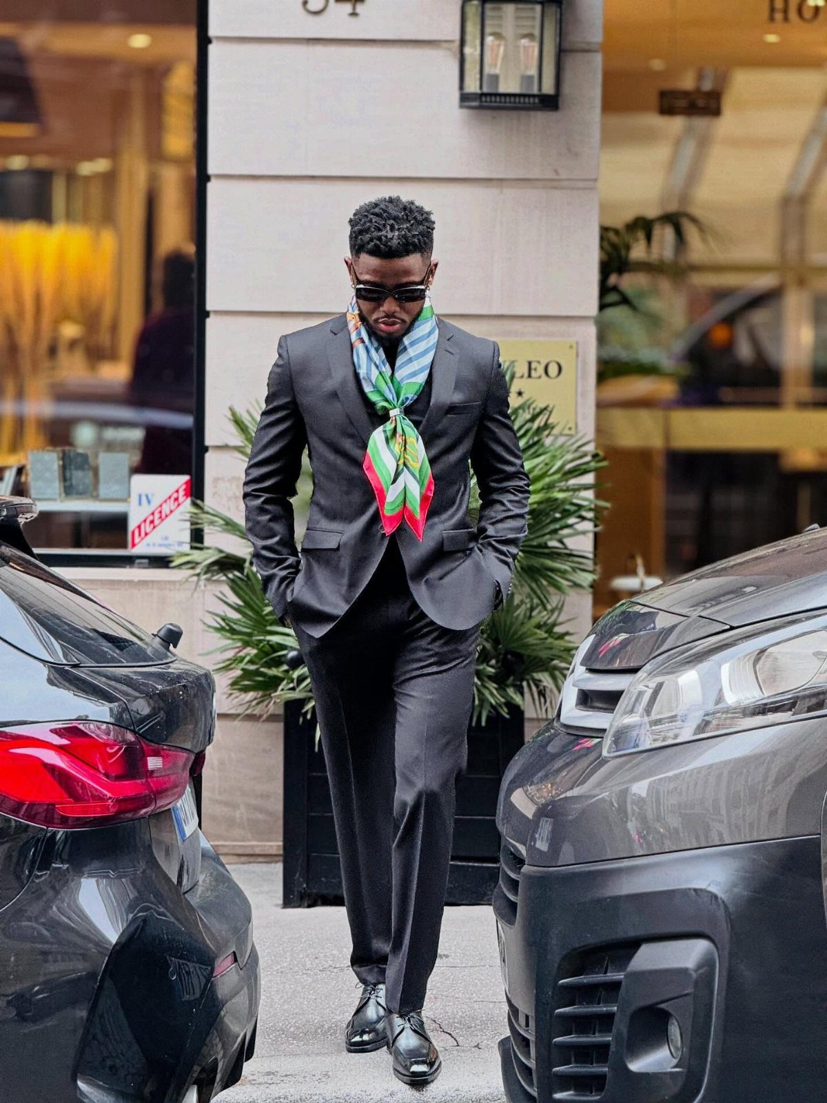 A man in a black blazer and trousers wearing a colorful patterned scarf on a city street.