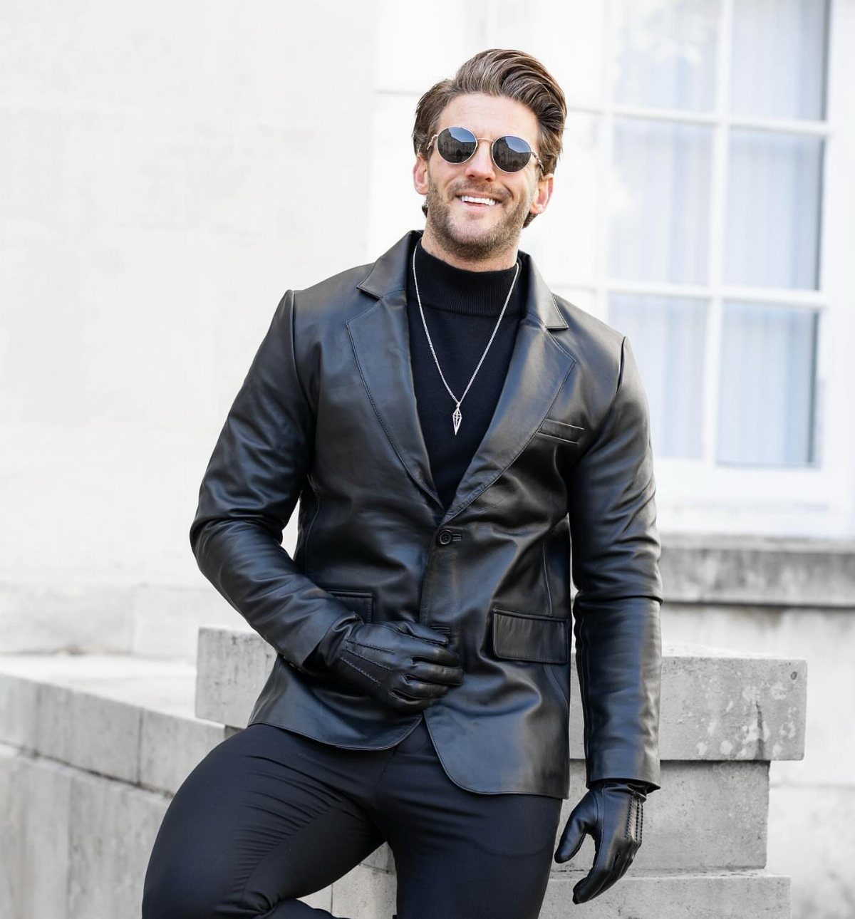 A man in a black leather blazer, black turtleneck, and black trousers smiling outdoors.