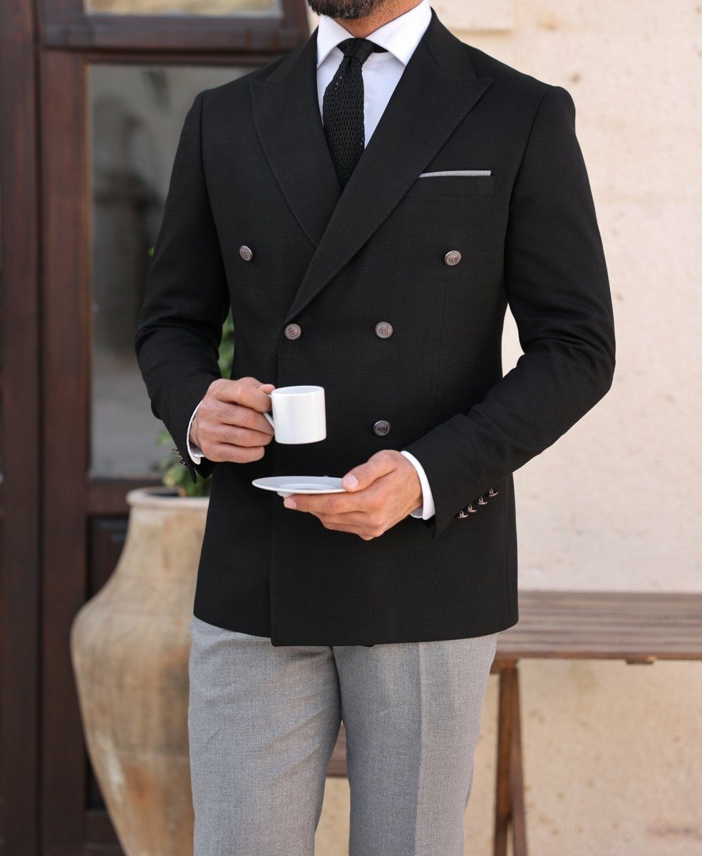 Man in a black double-breasted blazer and grey trousers holding a white coffee cup.