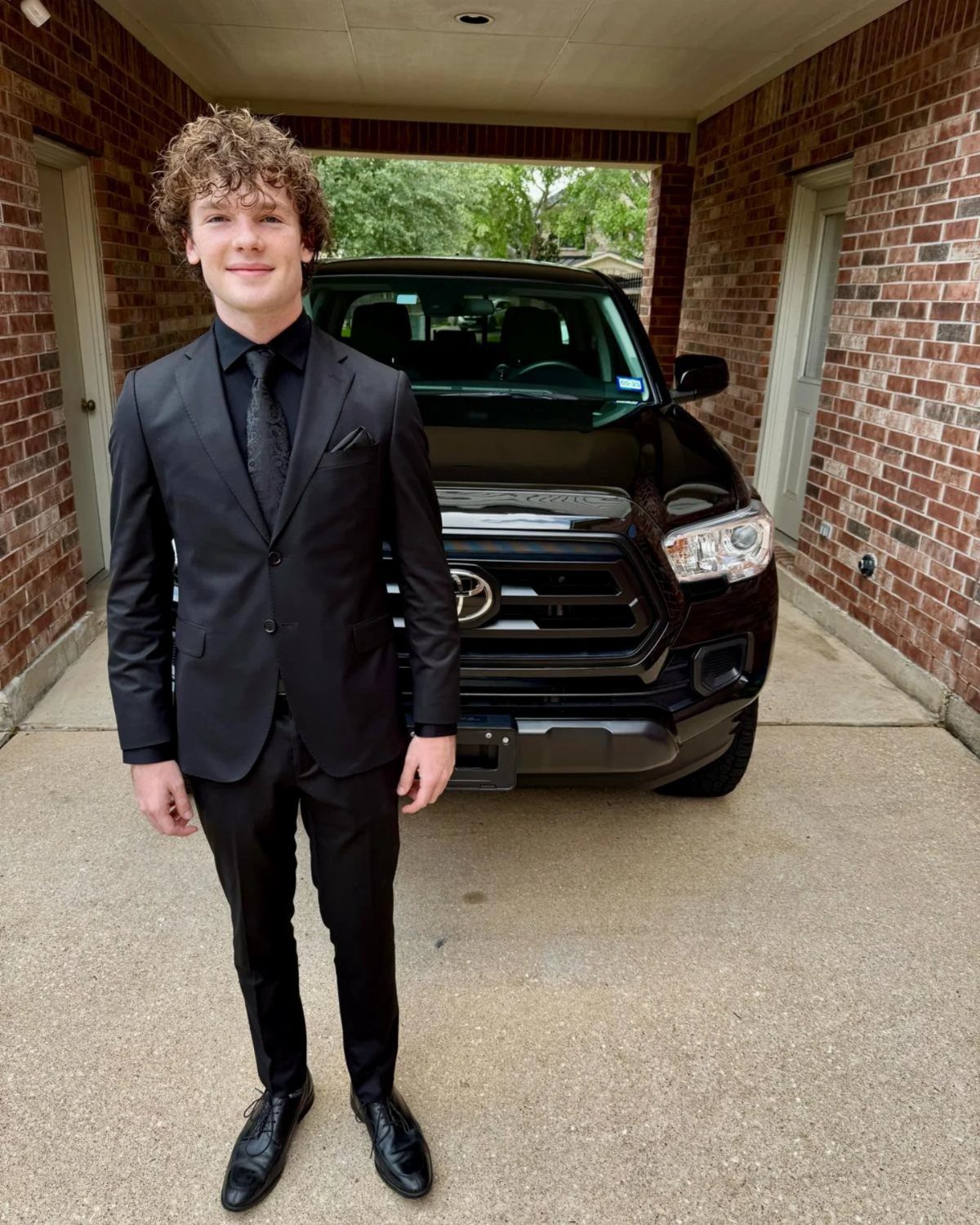 A young man smiling while wearing an all-black suit and tie standing in front of a black truck.