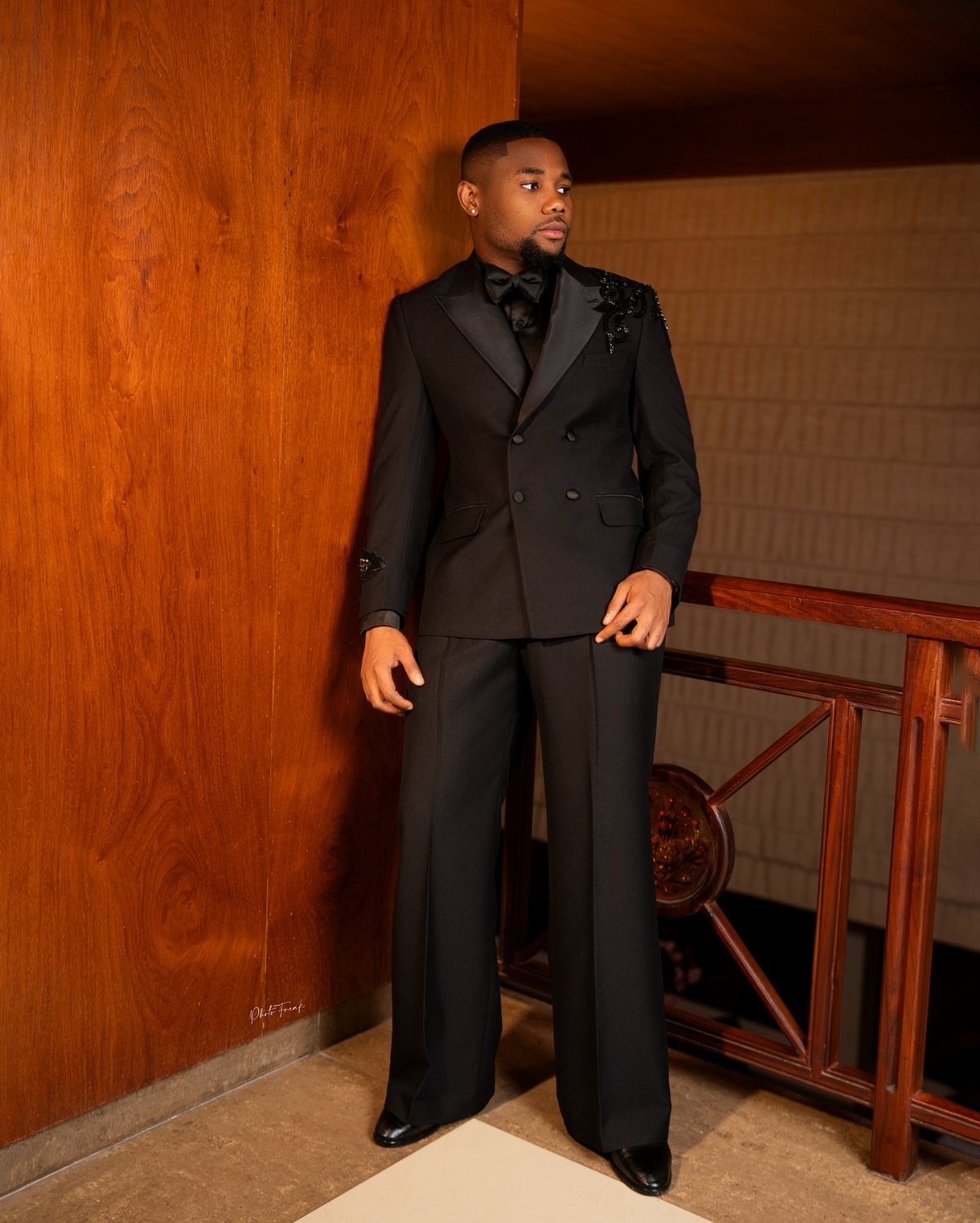 A man posing in a black double-breasted suit and bow tie against a wooden wall.