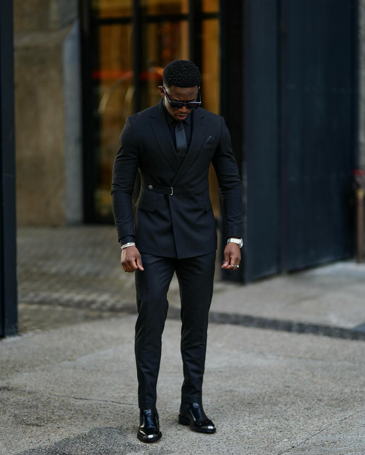 A man in a black double-breasted suit, black shirt, and tie posing outdoors.