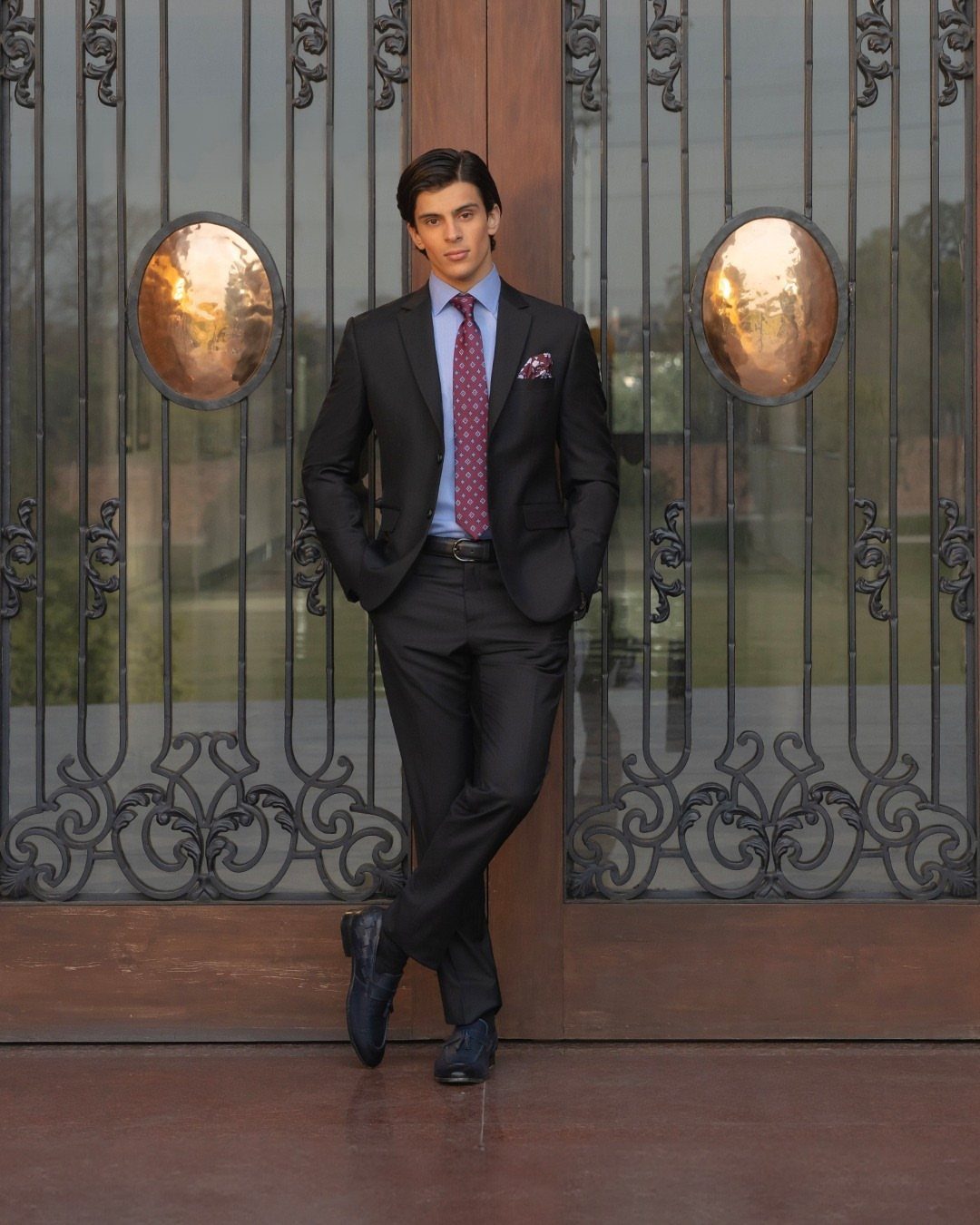 A man wearing a black suit, blue shirt, and red tie standing before a metal gate.