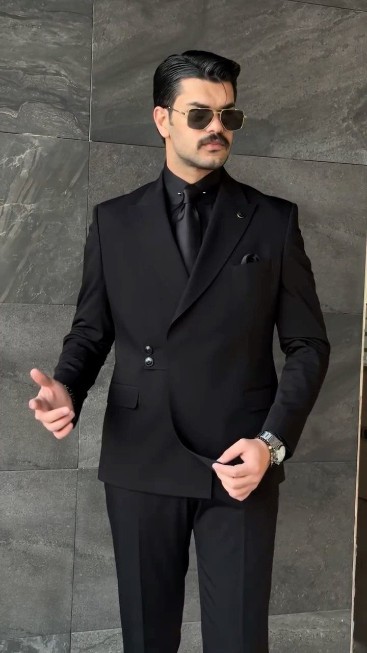 Man in a black suit and black tie wearing sunglasses against a grey stone wall.