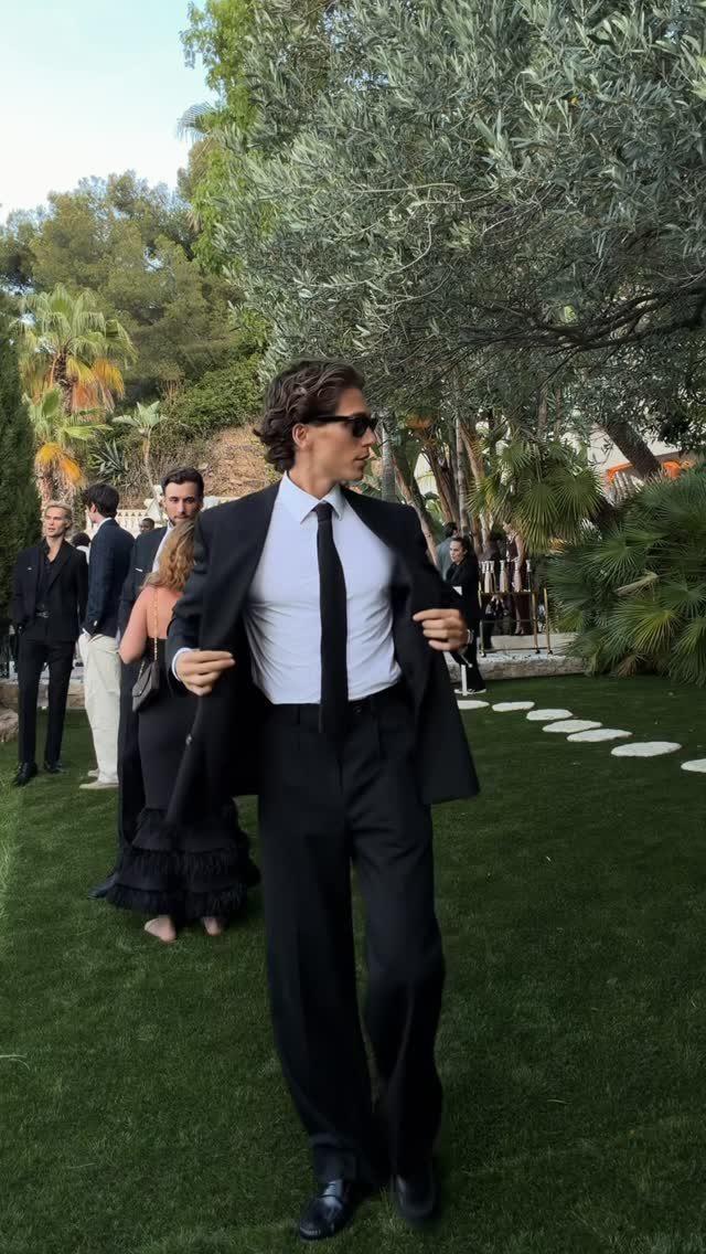 Man in a black suit, white shirt, and black tie standing on a green lawn.