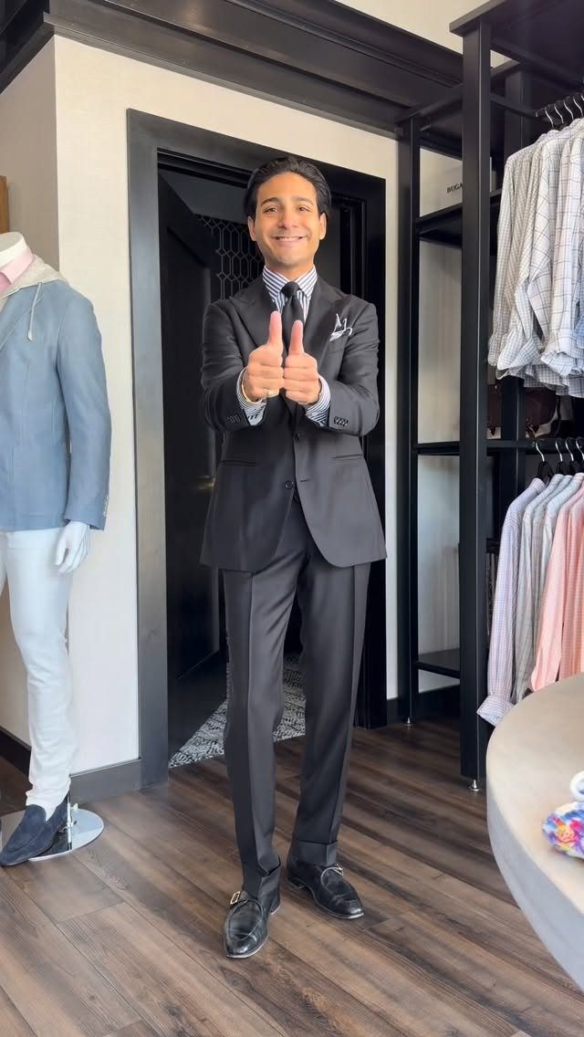 Smiling man in a black suit and black tie giving two thumbs up in a store.