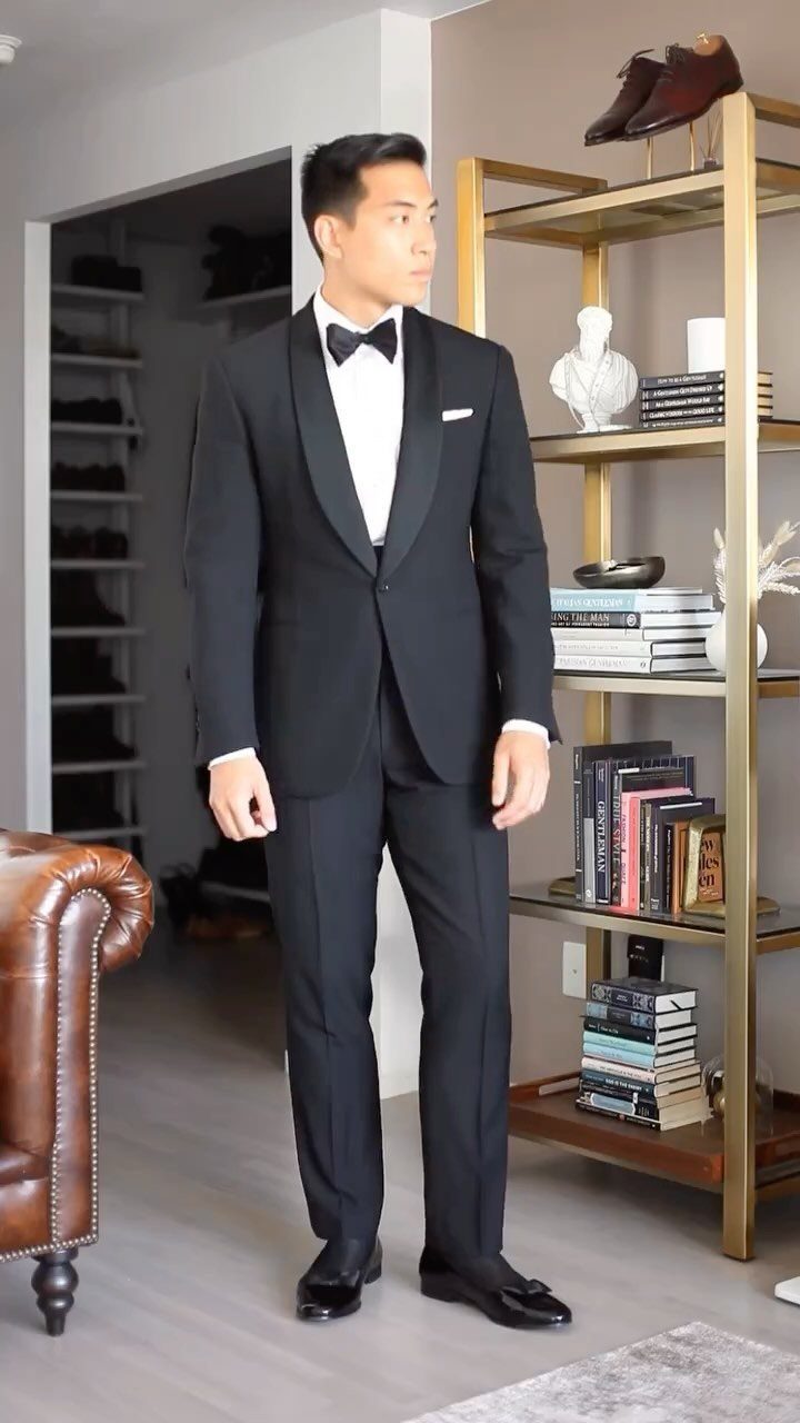 A man posing in a black shawl collar tuxedo with a white shirt and black bow tie.