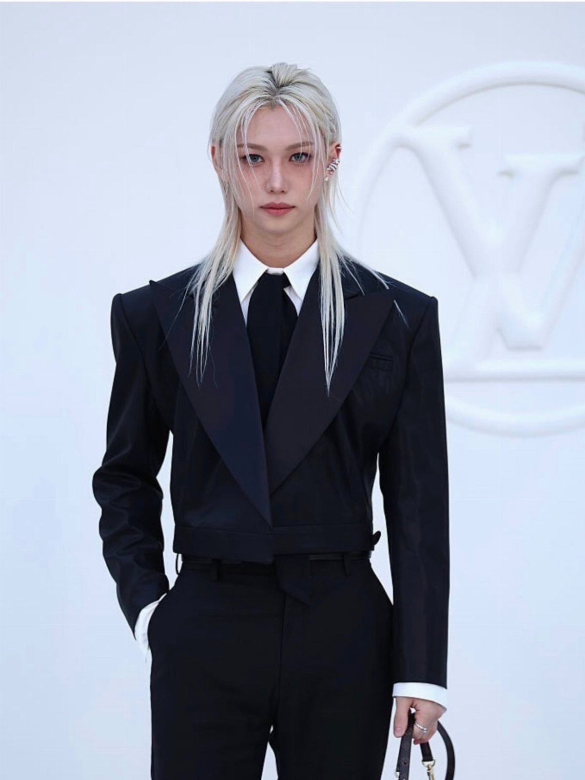 A person with long blonde hair wearing a black cropped tuxedo jacket and black tie.