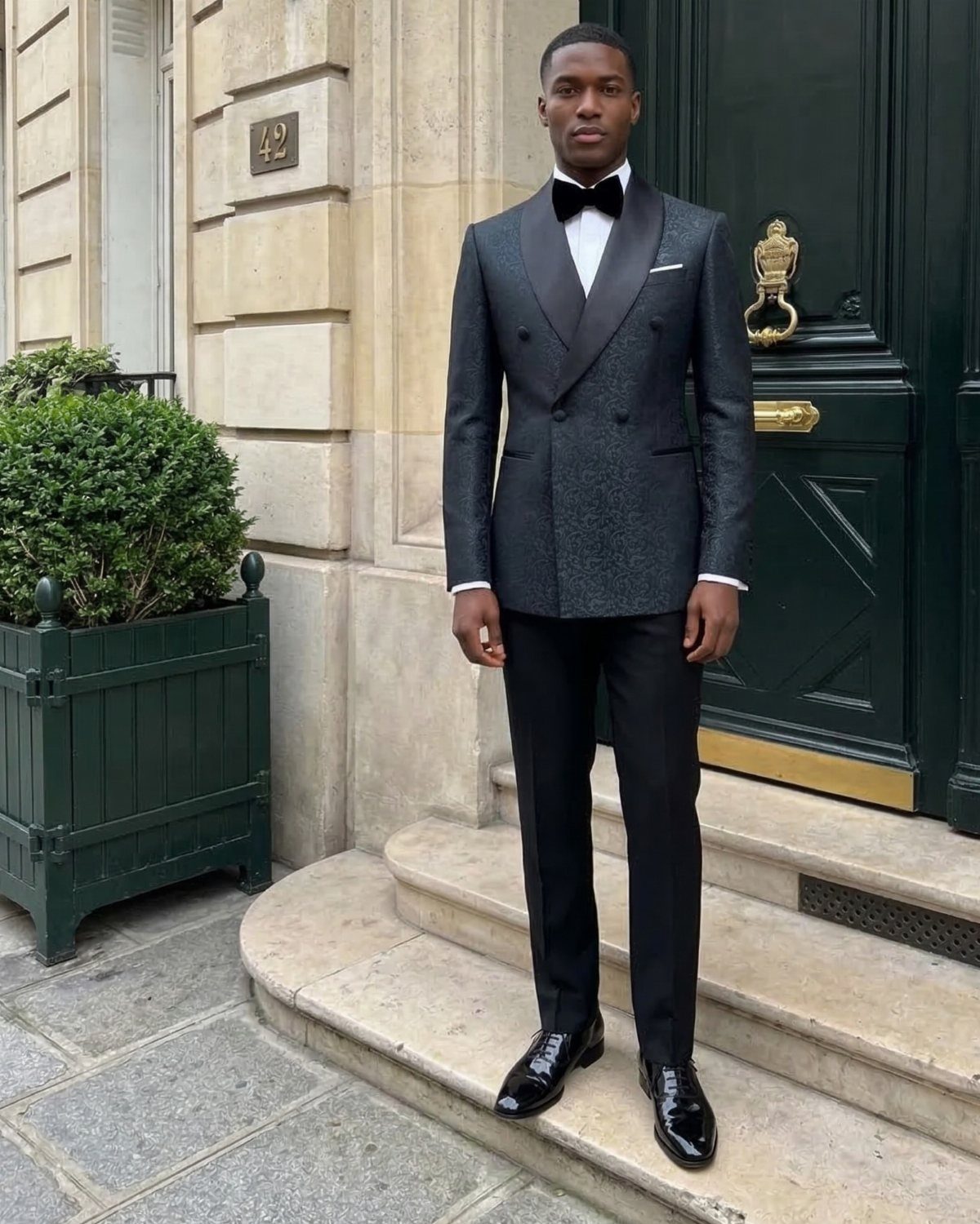 A man wearing a black double-breasted tuxedo and bow tie standing on stone steps.