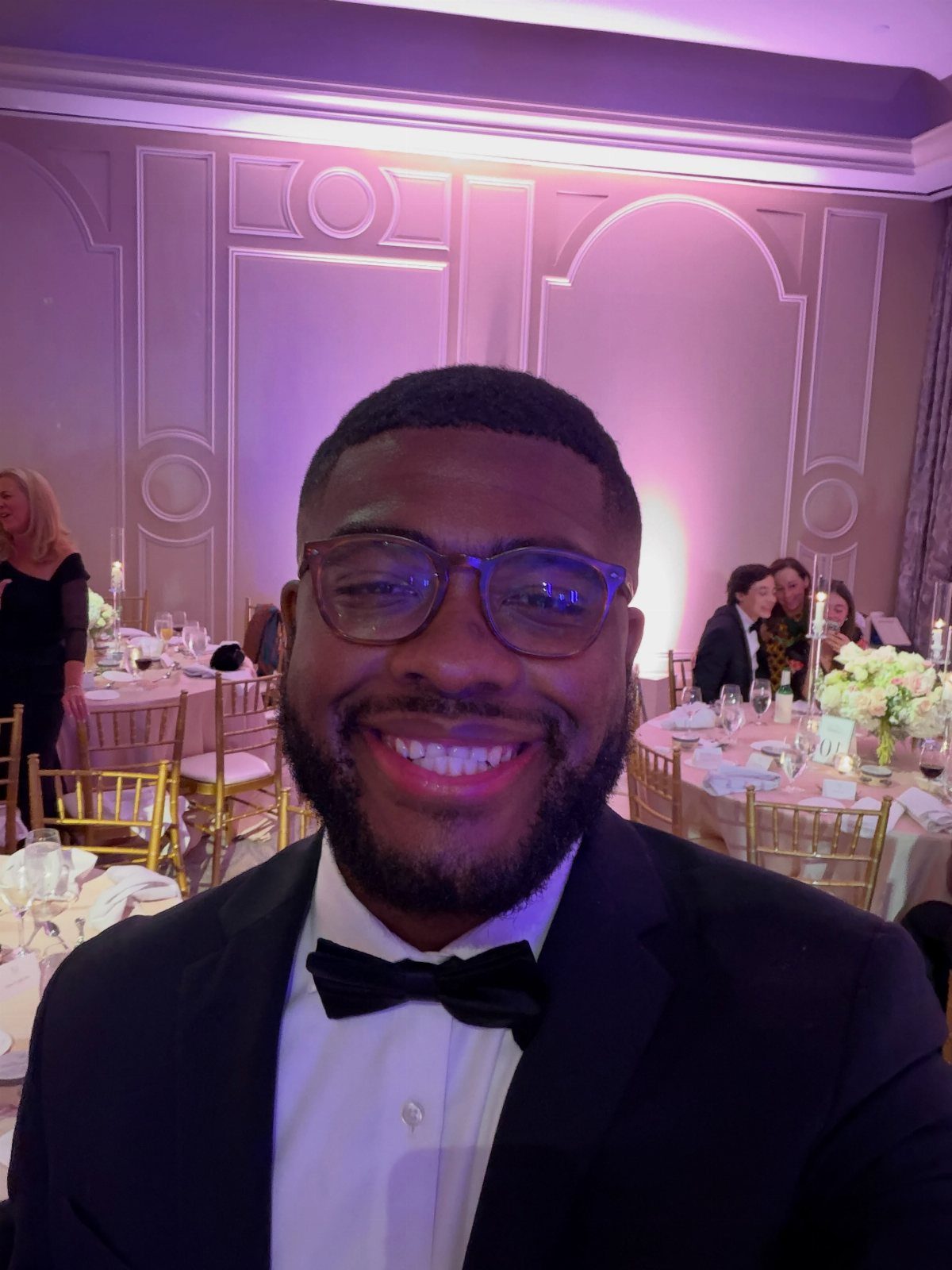 A smiling man wearing a black suit and a solid black bow tie at a formal event.