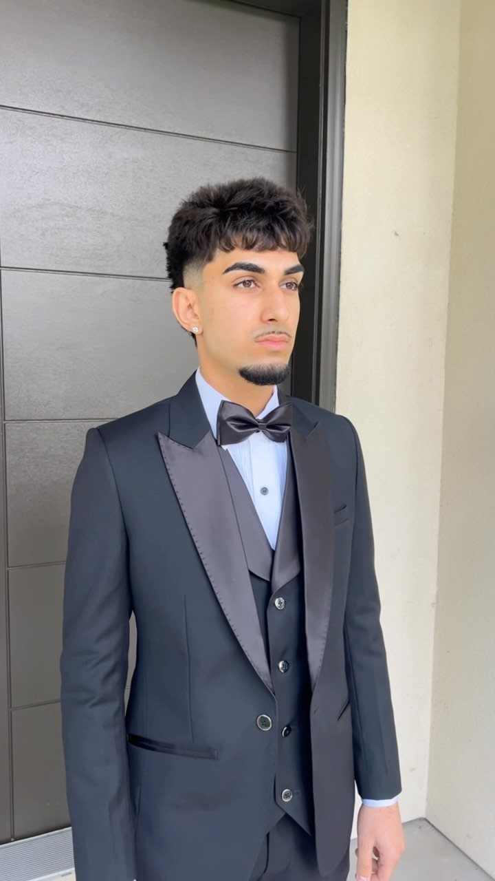 A young man posing in a formal black tuxedo with a bow tie and waistcoat.