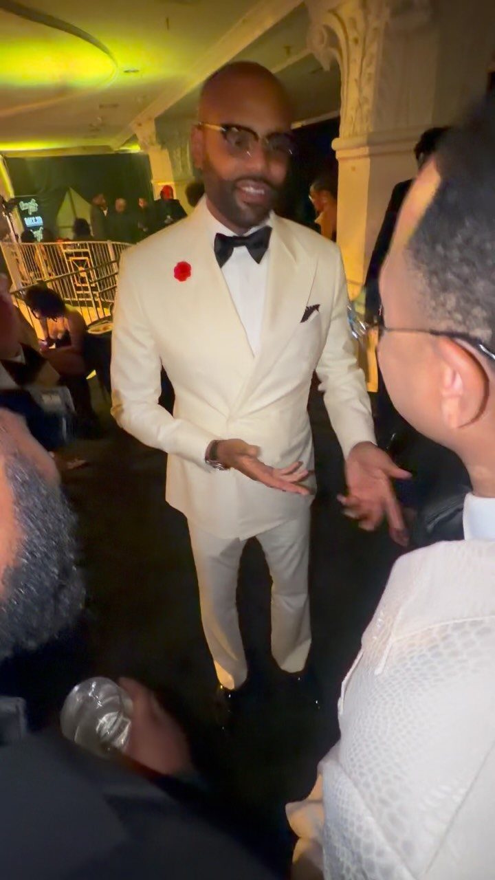 A man wearing a white tuxedo and black bow tie gestures during a formal event.