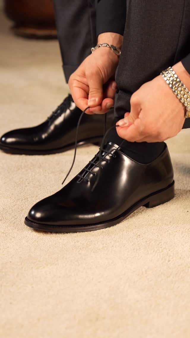 A person tying the laces of shiny black patent leather oxford shoes on a carpet.