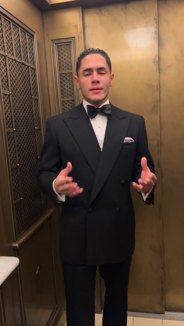 Man in a black double-breasted tuxedo and bow tie standing in a gold paneled hallway.