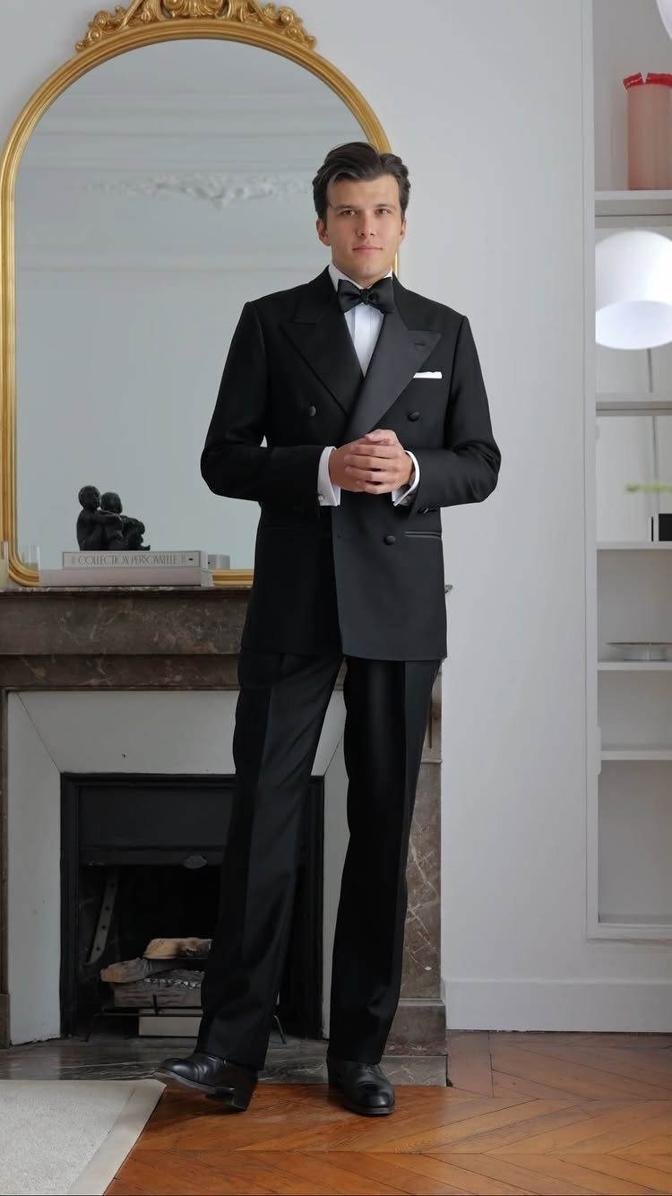 Man wearing a black double-breasted tuxedo and bow tie standing in a room.
