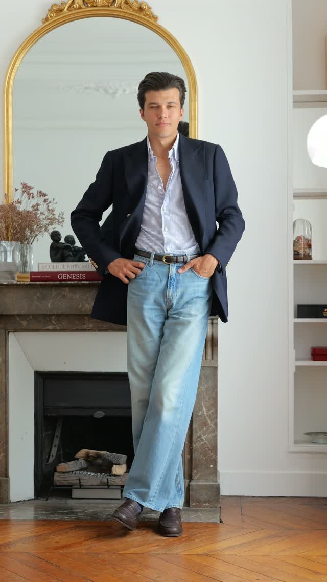 Man wearing a navy blazer, striped shirt, and light blue straight-leg jeans leaning on a fireplace.