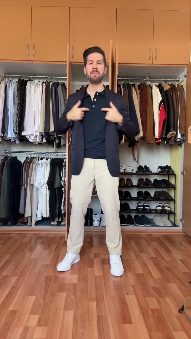 Man wearing a navy blazer, black polo, beige pants, and white sneakers standing in a closet.
