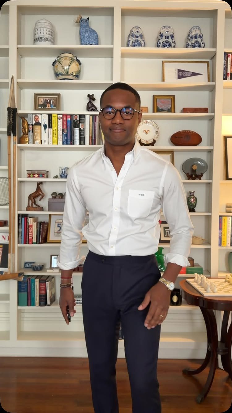 A man in a white button-down shirt and navy trousers standing before a bookshelf.