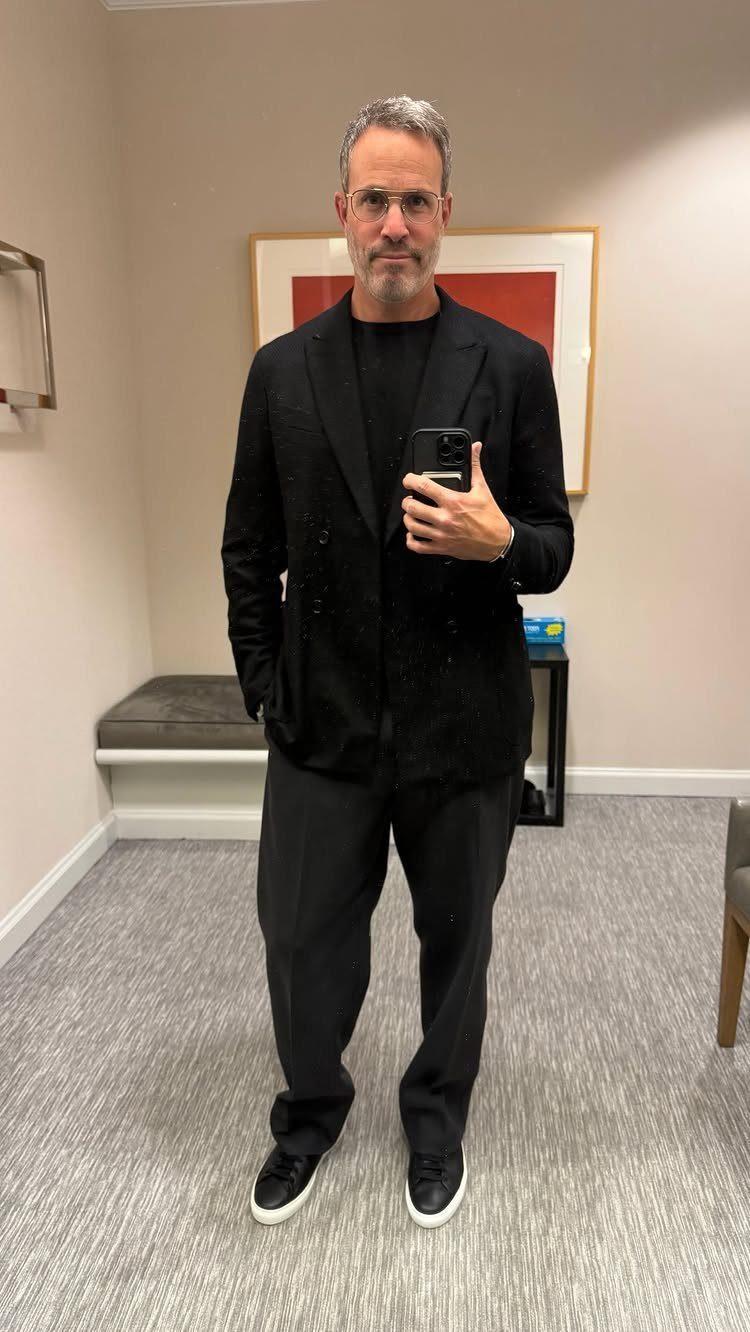 Man in an all-black outfit featuring a blazer, t-shirt, trousers, and sneakers.