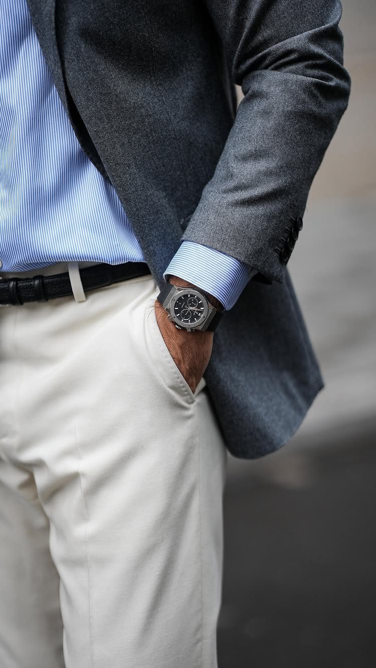 A man dressed in a grey blazer, blue striped shirt, and cream trousers with a black belt.