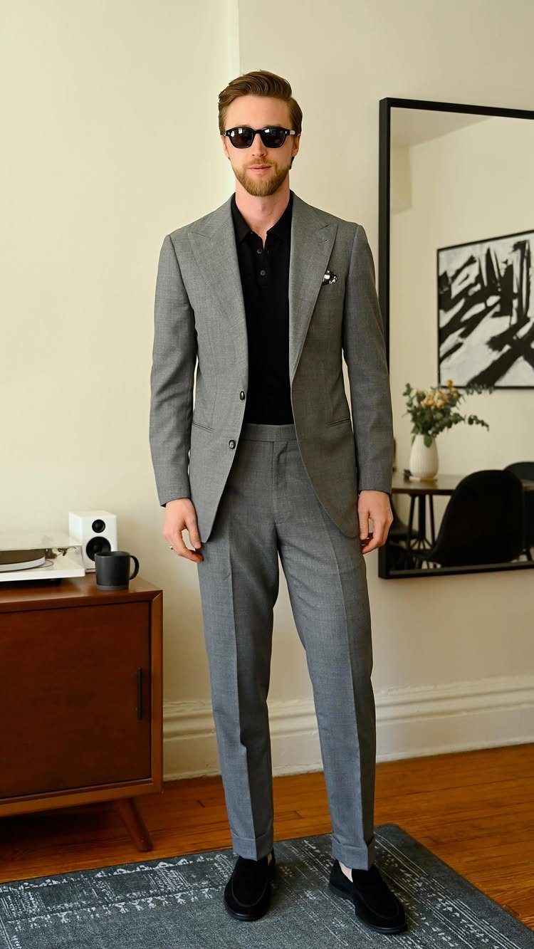 A man posing in a grey suit, black polo shirt, and sunglasses in a bright room.