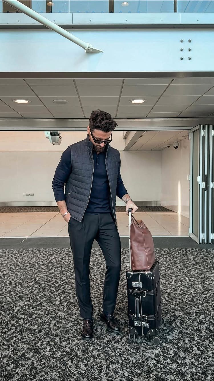 Man in business casual attire standing with a leather duffel bag and rolling suitcase in an airport.