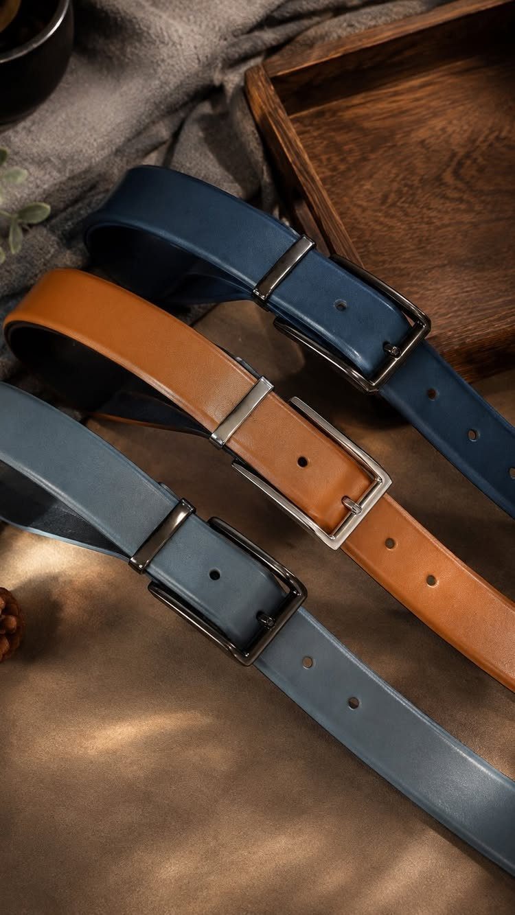 Three leather belts in navy, tan, and grey with silver buckles laid on a textured surface.