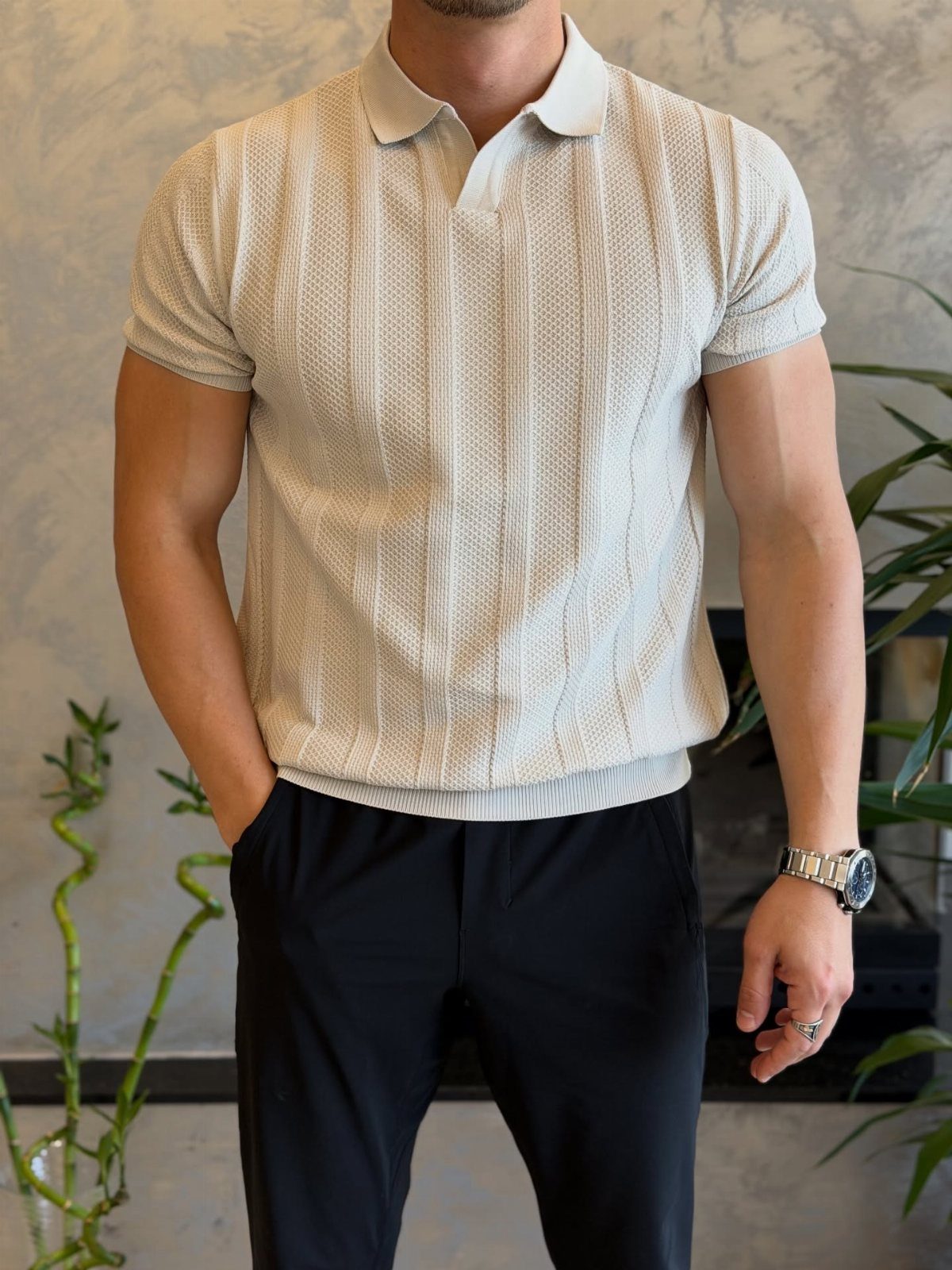 Man wearing a textured cream polo shirt and black trousers standing against a neutral wall.