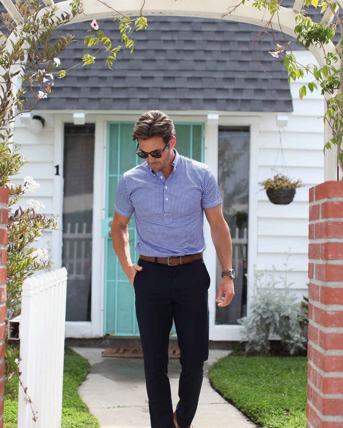 Man in a checkered shirt and navy trousers walking toward the camera outdoors.
