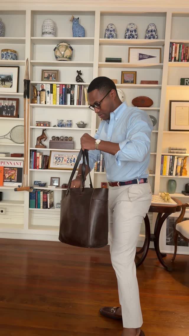 A man in a light blue shirt and beige trousers holds a large brown leather bag.