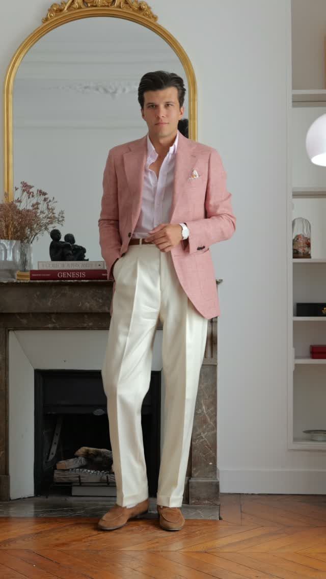 Man wearing a pink blazer, white collared shirt, and cream trousers standing by a fireplace.