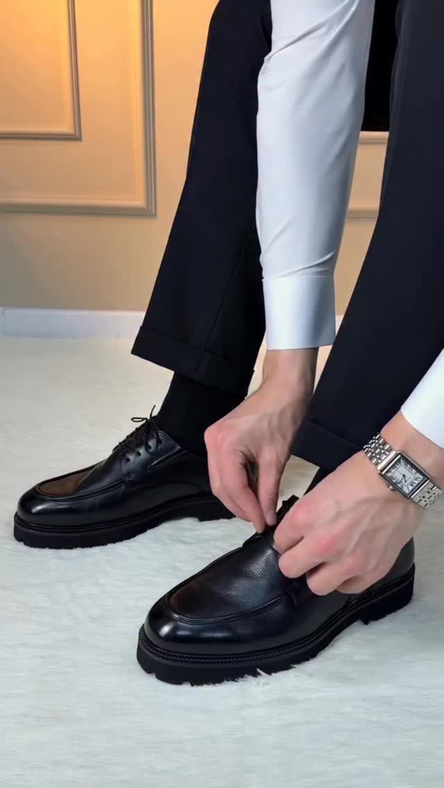 A person putting on black leather loafers with thick soles while wearing black trousers.