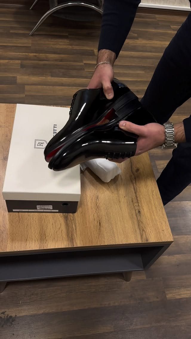 Hands holding a pair of polished black patent leather loafers over a white shoebox.