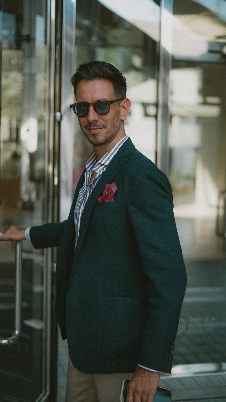 Man in a green blazer, striped shirt, and tan trousers standing by a glass door.