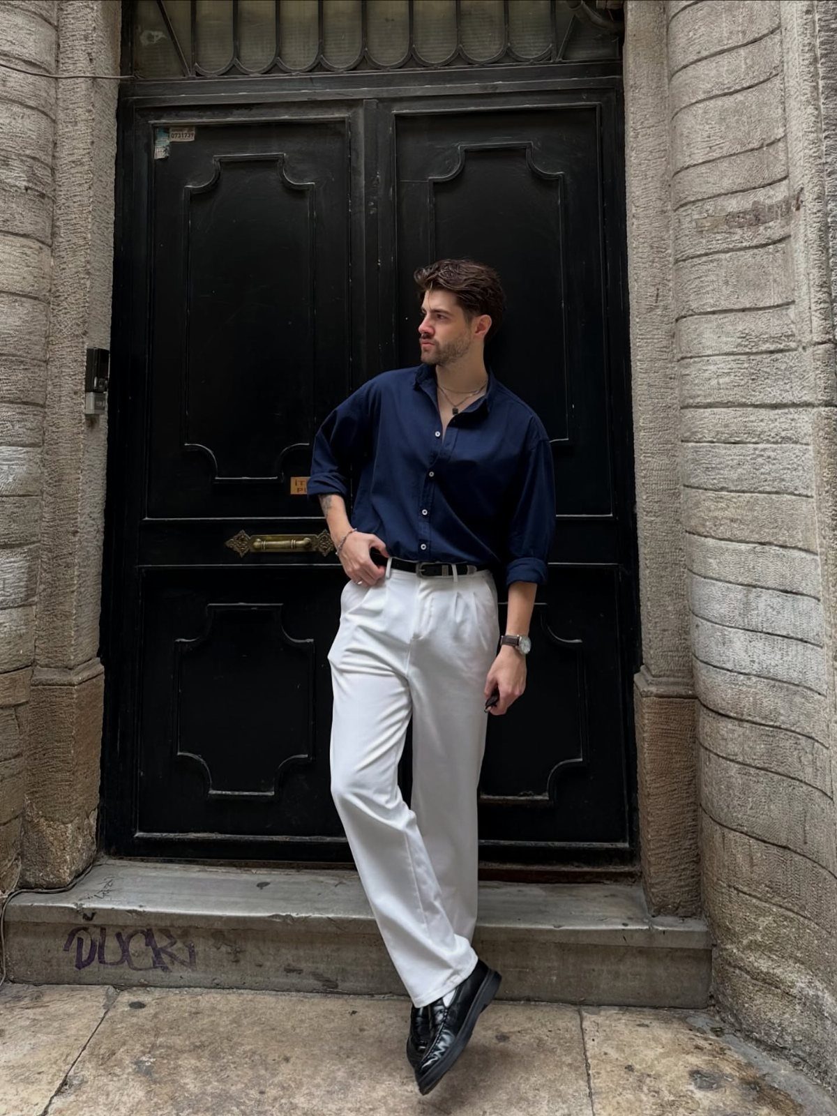 A man posing in a navy shirt and white trousers against a black door.