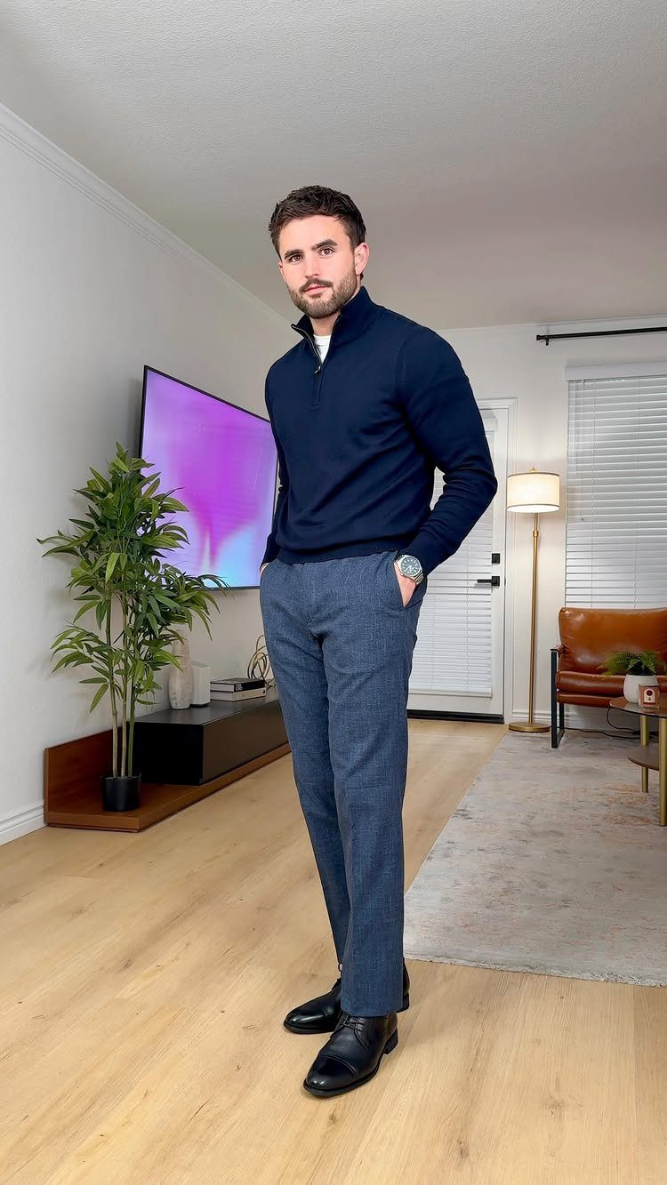 A man posing in a navy sweater and grey tailored trousers in a living room.
