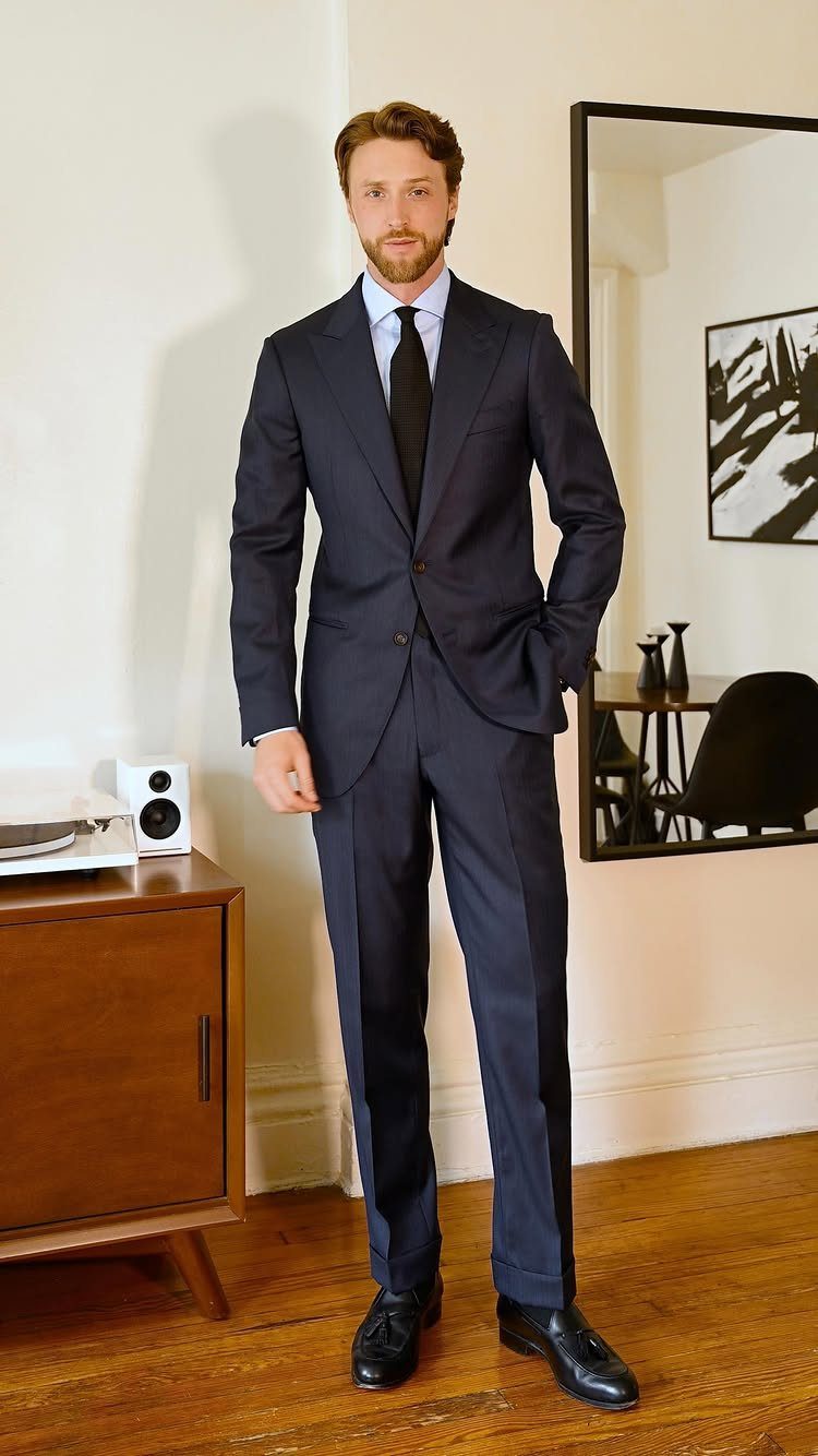 Man in a navy blue tailored suit and black tie standing in a room.
