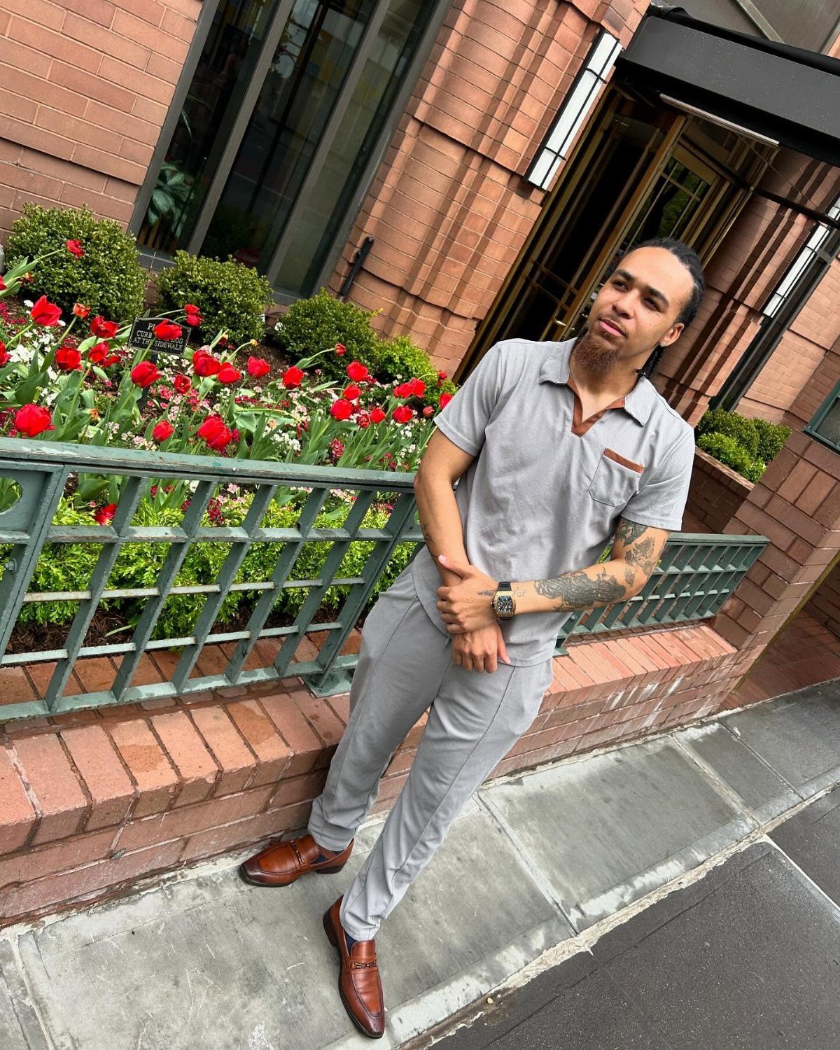A man posing in a grey polo shirt, grey trousers, and brown leather loafers outdoors.